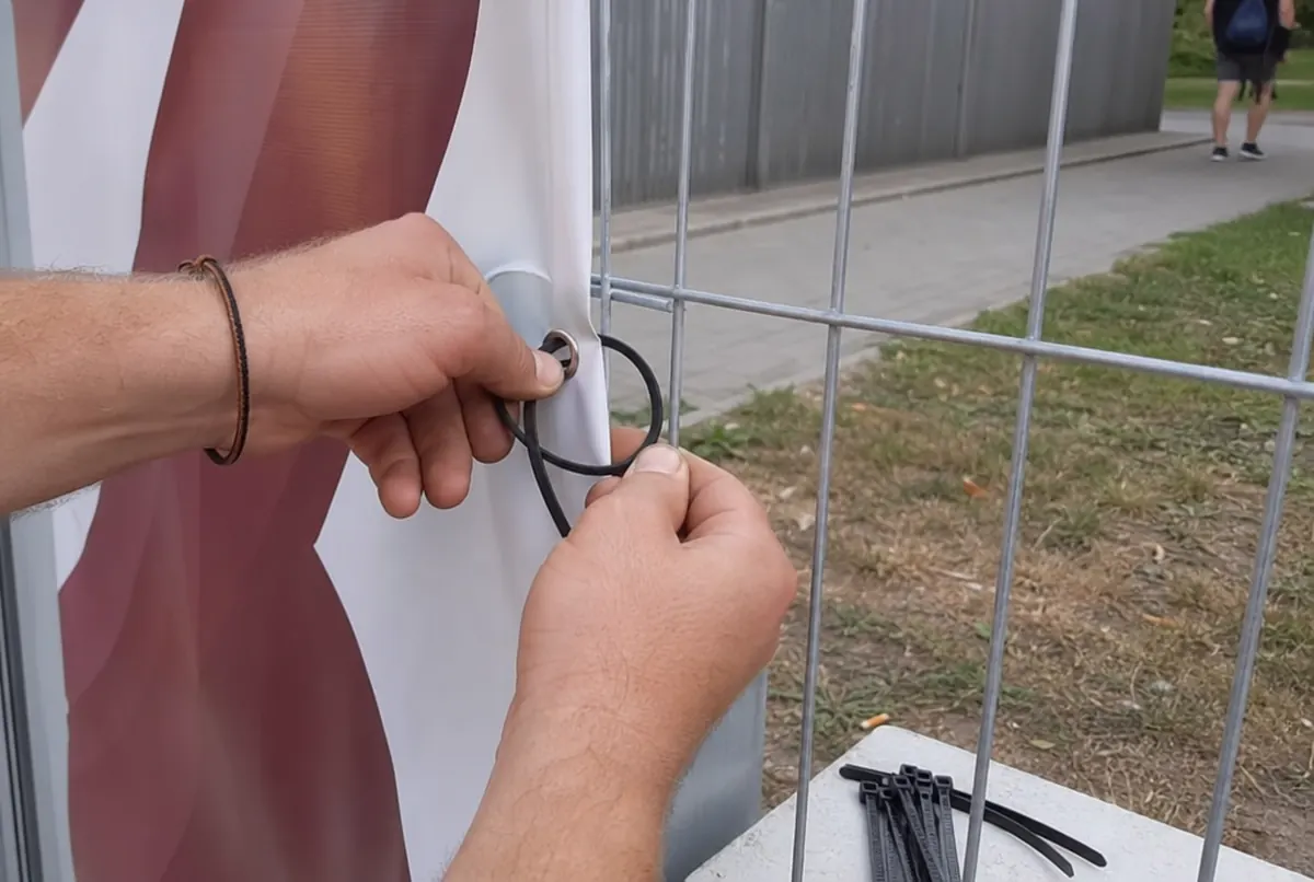 Ręce mocują baner gumą przez oczko do panelu ogrodzenia