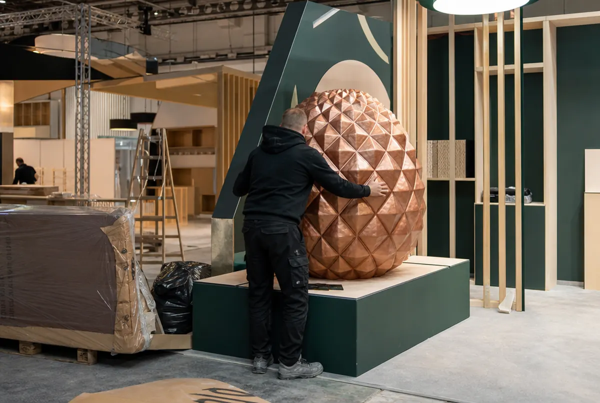 A large copper-painted 3D printed element being placed on a trade fair stand plinth