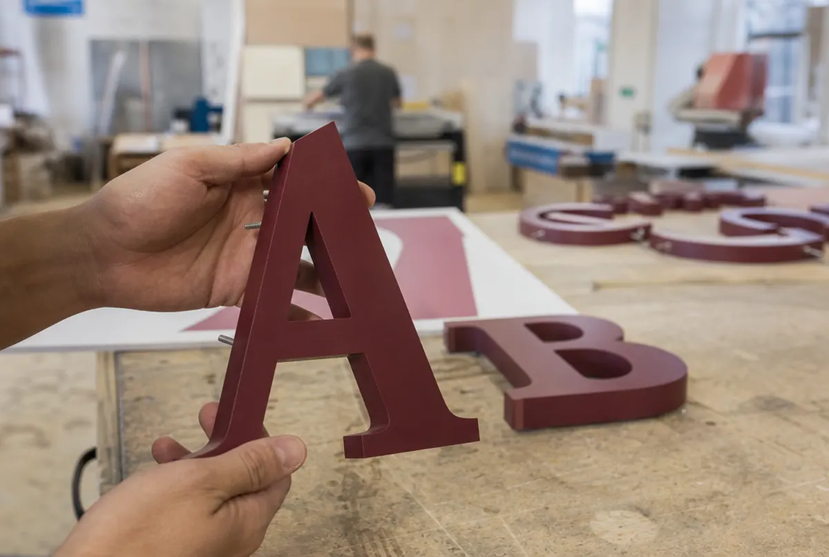 Manos sostienen una letra 3D burdeos de PVC con canto fresado y pernos de montaje
