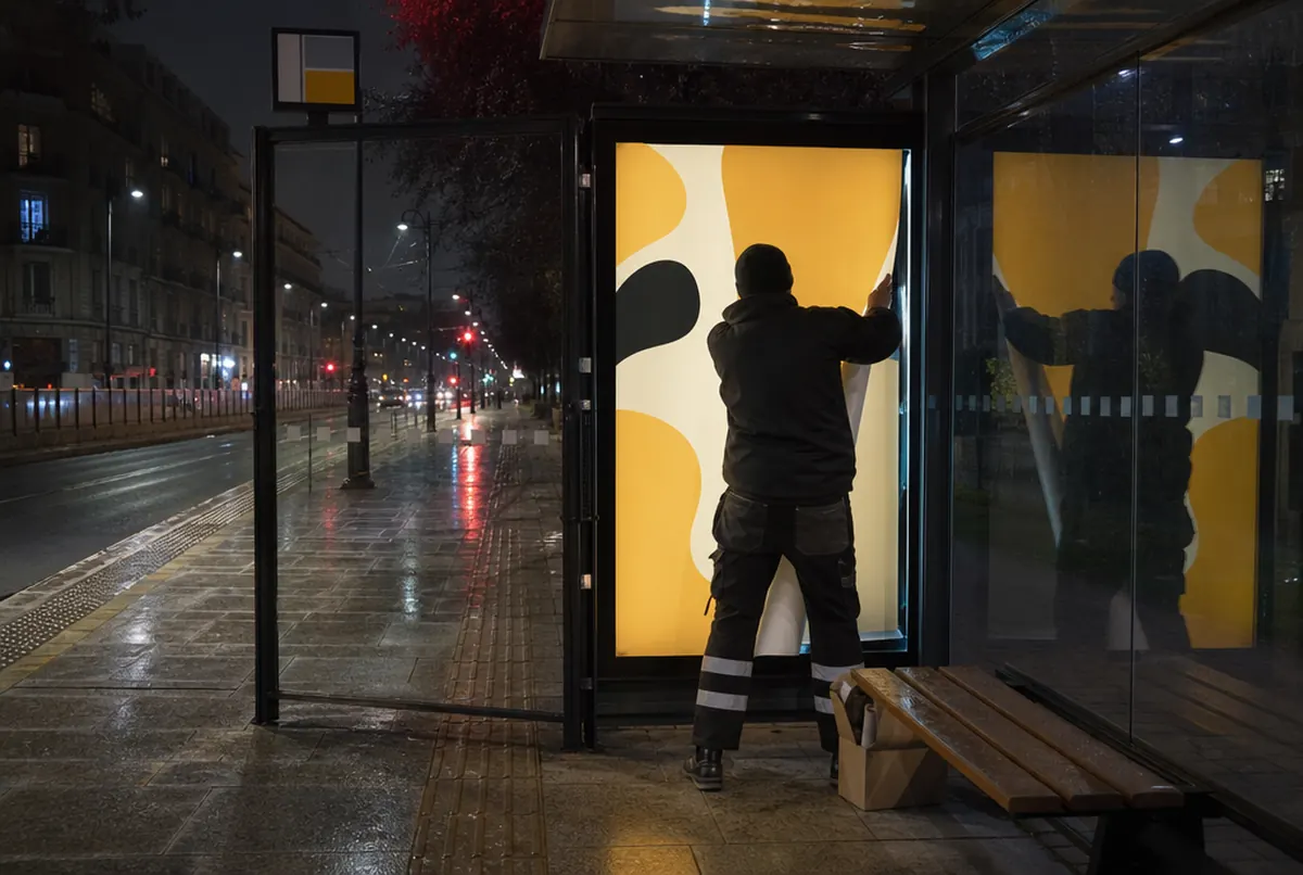 Un agent change l'affiche d'une vitrine citylight éclairée sous un abribus la nuit